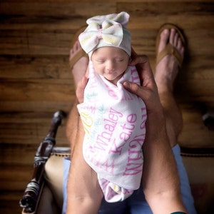 Puede incluir: Una reci&eacute;n nacida con un gorro de punto rosa y blanco con un lazo, envuelta en una manta blanca con letras rosas y verdes que dicen "Whaley Kate" y "Made with Love".