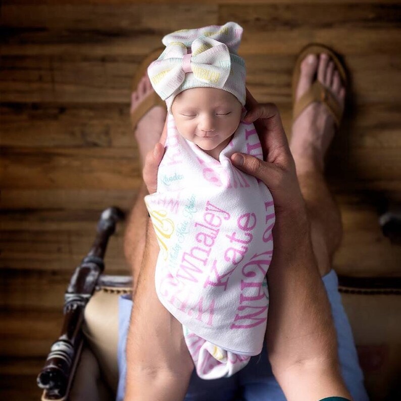 Puede incluir: Una reci&eacute;n nacida con un gorro de punto rosa y blanco con un lazo, envuelta en una manta blanca con letras rosas y verdes que dicen "Whaley Kate" y "Made with Love".