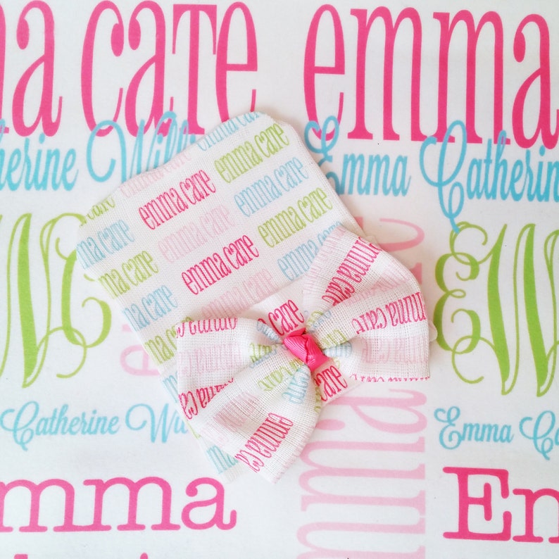 Puede incluir: Un gorro blanco para beb&eacute; con un lazo rosa. El gorro est&aacute; cubierto con un patr&oacute;n repetido de los nombres "Emma Care" en rosa, azul y verde.