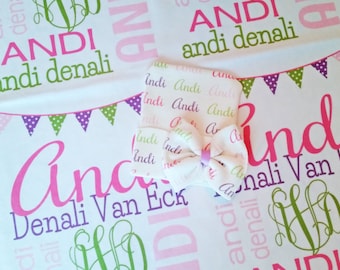 May include: A white baby blanket with a pink and green pattern featuring the name "Andi Denali" repeated throughout. A small white burp cloth with a pink bow is on top of the blanket.