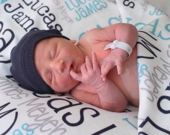 May include: A newborn baby wearing a navy blue beanie lies on a blanket. The blanket is white with the name "Lucas James Meyer" printed in black and light blue. The baby has a white hospital band on its wrist.