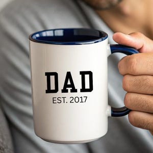 May include: White ceramic mug with a navy blue interior and handle. The mug features the word "DAD" in bold black letters, with "EST. 2017" below. The mug is held by a person.