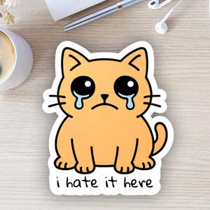 以下が含まれることがあります： オレンジ色の毛並み、黒い目、涙を持つ漫画の猫のステッカー。猫は悲しい表情をしており、下に「i hate it here」というテキストがあります。