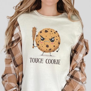 May include: A white long-sleeved t-shirt featuring a cartoon cookie holding a baseball bat. The cookie has chocolate chips and a grumpy expression. The text "Tough Cookie" is printed below the cookie.