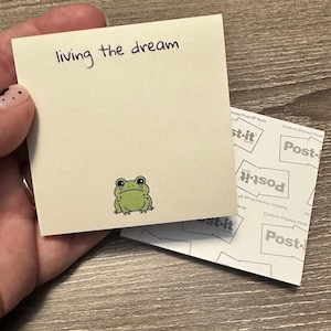 May include: A cream-colored Post-it Note with the handwritten phrase "living the dream" and a small green frog illustration with tear drops. The note is partially covering a Post-it pad.