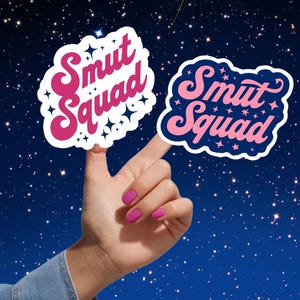 以下が含まれることがあります： レトロなフォントで「Smut Squad」というテキストが入った2つのステッカー。1つはピンクのテキストと星が付いた白い縁取り、もう1つはピンクのテキストと星が付いた濃い青色の縁取りです。背景は濃い青色の星空です。