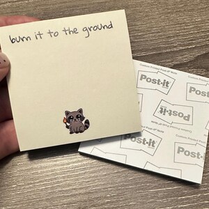 May include: A beige Post-it note with the handwritten phrase "burn it to the ground." A cartoon raccoon holding a lit match is printed on the note. The Post-it note is next to a stack of Post-it notes.