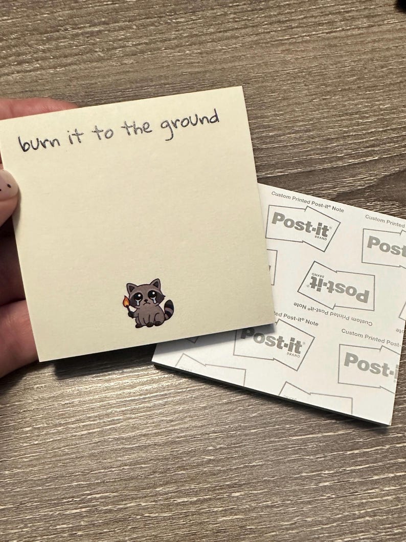 May include: A beige Post-it note with the handwritten phrase "burn it to the ground." A cartoon raccoon holding a lit match is printed on the note. The Post-it note is next to a stack of Post-it notes.