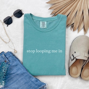 以下が含まれることがあります： 白い文字で「stop looping me in」とプリントされたターコイズブルーのTシャツ。白い背景に、サングラス、ゴールドのチェーンネックレス、ベージュのクロッグと一緒に置かれています。