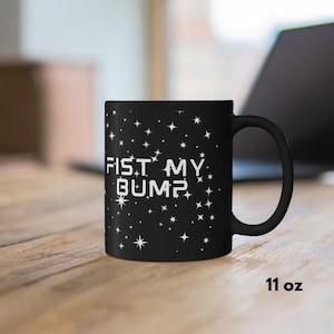 May include: A white ceramic mug with a black handle and the text "But First, Coffee" in a bold, black font. The mug is set against a blurred background, suggesting a cozy setting.