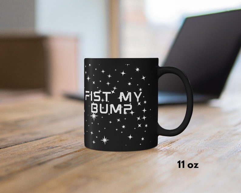 May include: A white ceramic mug with a black handle and the text "But First, Coffee" in a bold, black font. The mug is set against a blurred background, suggesting a cozy setting.