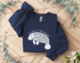 May include: A navy blue sweatshirt featuring a white manatee design with swirling patterns. The text "RESPECT THE LOCALS" is above the manatee. The sweatshirt is folded on a wooden surface with floral accents.