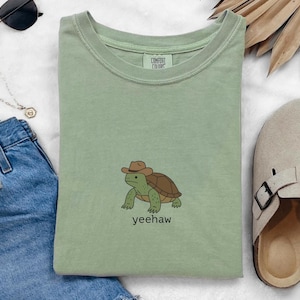 May include: Sage green t-shirt featuring a cartoon turtle wearing a cowboy hat, with the word "yeehaw" below. The shirt is folded, with a pair of sunglasses, a necklace, and denim shorts visible in the background.