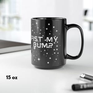 May include: Black 15 oz coffee mug with the text "FIST MY BUMP" in white, surrounded by white stars. The mug has a large handle and is on a white surface.