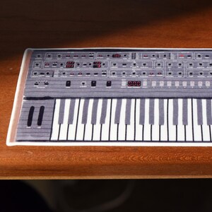 Könnte beinhalten: Rechteckige, grau-weiße Matte mit einem Keyboard-Design und dem Text "Prophet-6". Die Matte zeigt eine detaillierte Illustration eines Synthesizers mit zahlreichen Knöpfen, Reglern und einer Tastatur mit schwarzen und weißen Tasten. Der Rand der Matte ist in Holzoptik gehalten.