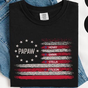 May include: Black t-shirt with a distressed American flag design. The flag includes the word "PAPAW" surrounded by stars, and the names Henry, Emma, Stella, and Colson. The shirt is displayed on a pair of blue jeans.