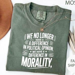 May include: Moss green t-shirt with the text "WE NO LONGER HAVE A DIFFERENCE IN POLITICAL OPINION. WE HAVE A DIFFERENCE IN MORALITY." A gold necklace and a brown hat are also visible.