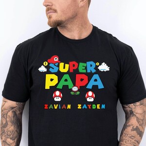 以下が含まれることがあります： ビデオゲーム風のカラフルな「Super Papa」グラフィックがプリントされた黒いTシャツ。赤い帽子、雲、キノコ、そして「Zavian」と「Zayden」の名前がデザインされています。お父さんへの楽しいギフトです。