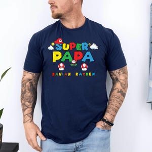 以下が含まれることがあります： カラフルなブロック文字で「SUPER PAPA」と書かれた、2つのキノコのグラフィックが特徴のネイビーブルーのTシャツ。テキストの下には、虹色のフォントで「ZAVIAN ZAYDEN」という名前があります。カジュアルなスタイルのシャツです。