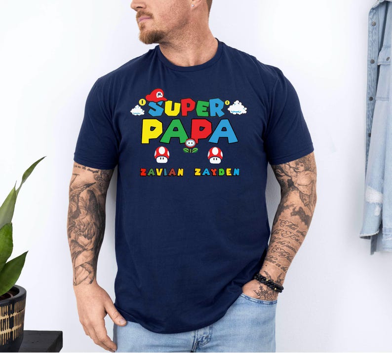 以下が含まれることがあります： カラフルなブロック文字で「SUPER PAPA」と書かれた、2つのキノコのグラフィックが特徴のネイビーブルーのTシャツ。テキストの下には、虹色のフォントで「ZAVIAN ZAYDEN」という名前があります。カジュアルなスタイルのシャツです。