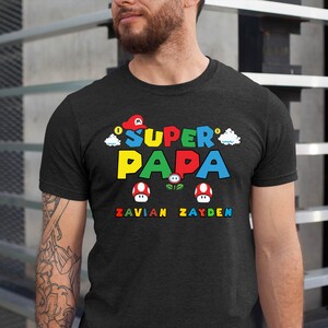 以下が含まれることがあります： 鮮やかな「Super Papa」のグラフィックがプリントされたダークグレーのTシャツ。マリオをテーマにしたデザインで、赤い帽子、雲、キノコが描かれています。「Zavian」と「Zayden」の名前が下に印刷されています。