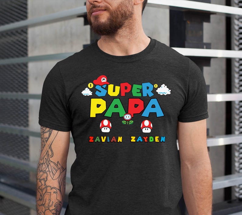 以下が含まれることがあります： 鮮やかな「Super Papa」のグラフィックがプリントされたダークグレーのTシャツ。マリオをテーマにしたデザインで、赤い帽子、雲、キノコが描かれています。「Zavian」と「Zayden」の名前が下に印刷されています。