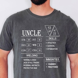 May include: A dark gray t-shirt with a white graphic design that resembles a character sheet from a role-playing game. The graphic includes the word "UNCLE" at the top, followed by stats like "STR 18", "INT 16", "DEX 10", "WIS 8", "CON 17", "CHA 14", "HP 53", "AC 15", and "SKILLS". The graphic also includes a list of skills and immunities.