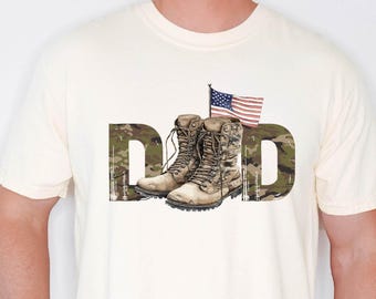May include: Cream-colored t-shirt with the word "DAD" in a camouflage pattern. The letter "D" is decorated with a pair of combat boots and an American flag. The shirt is a casual style.
