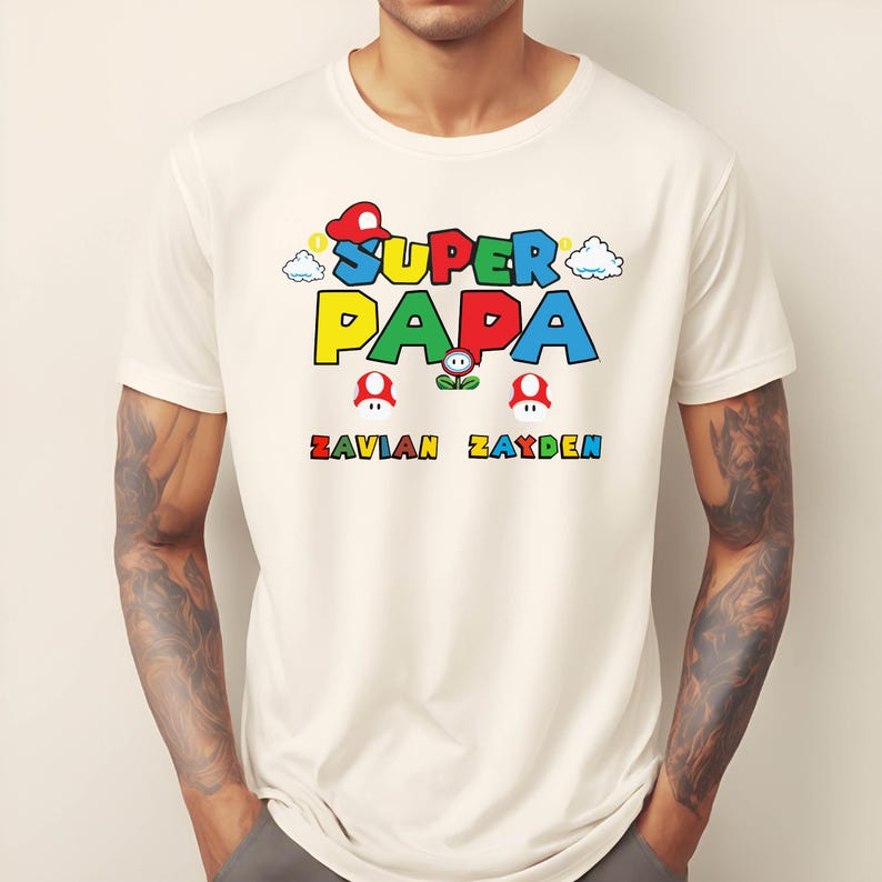 以下が含まれることがあります： クリーム色のTシャツ。カラフルなグラフィックで「SUPER PAPA」と遊び心のあるフォントで書かれています。テキストの下には、同じフォントで「ZAVIAN ZAYDEN」という名前があります。デザインにはキノコと雲のグラフィックが含まれています。