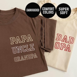 May include: Two t-shirts with embroidered text. The brown t-shirt says "Papa Uncle Grandpa" in white text. The beige t-shirt says "Dad Opa" in red text.