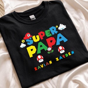 以下が含まれることがあります： カラフルなブロック文字で「SUPER PAPA」と書かれた黒いTシャツ。赤い帽子、キノコ、雲の漫画グラフィックが特徴です。「ZAVIAN ZAYDEN」という名前が下に印刷されています。