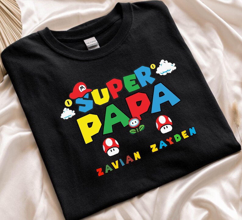 以下が含まれることがあります： カラフルなブロック文字で「SUPER PAPA」と書かれた黒いTシャツ。赤い帽子、キノコ、雲の漫画グラフィックが特徴です。「ZAVIAN ZAYDEN」という名前が下に印刷されています。