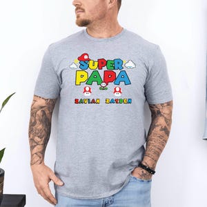 以下が含まれることがあります： 「SUPER PAPA」と書かれたカラフルなグラフィックが特徴のライトグレーのTシャツ。テキストの下には、緑色の「SAVIAN ZAYDEN」という名前があります。デザインにはキノコと雲のグラフィックが含まれています。
