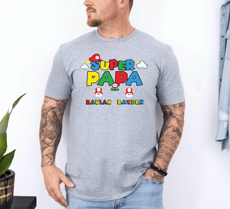 以下が含まれることがあります： 「SUPER PAPA」と書かれたカラフルなグラフィックが特徴のライトグレーのTシャツ。テキストの下には、緑色の「SAVIAN ZAYDEN」という名前があります。デザインにはキノコと雲のグラフィックが含まれています。