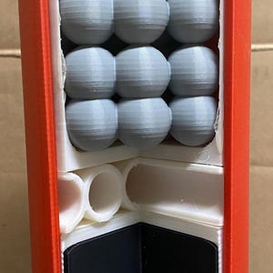 May include: A red and yellow plastic container with a white interior. The container is filled with gray balls. The container is open and the balls are visible. The container has a black and white base.
