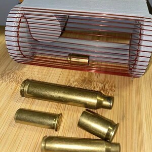 May include: A collection of brass bullet casings of varying sizes, some with spent primers, are arranged on a wooden surface. A rectangular object with copper-colored wire and a gray top is in the background.