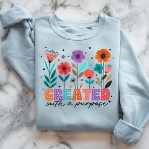 以下が含まれることがあります： 明るいブルーのトレーナーに、カラフルな花柄と「CREATED with a purpose」の文字がプリントされています。