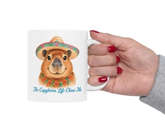 Op de afbeelding: Witte keramische mok met een cartoon capybara die een sombrero en een bloemenkrans draagt. De tekst "The Capybara Life Chose Me" staat op de mok gedrukt.