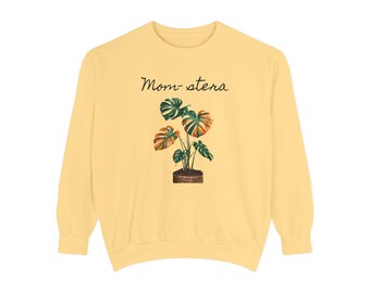 May include: A yellow sweatshirt with the text "Mom-stera" printed on the front. The text is in black and is above a graphic of a Monstera plant in a brown pot.