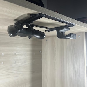 Può includere: Un supporto nero sotto la scrivania per un controller per videogiochi e un auricolare. Il supporto è fissato alla parte inferiore di una scrivania con viti. Il controller è appeso al supporto e l'auricolare è appeso a un gancio sul supporto.