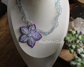 May include: A multi-strand necklace featuring a large, layered felt flower pendant in shades of purple and light blue. The flower is embellished with silver and white beads. The necklace chain consists of delicate, twisted strands of light blue and pink seed beads.