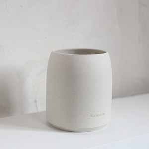 Puede incluir: Un jarrón de cerámica blanco roto moteado, de forma redondeada y con una abertura ancha. La palabra "Karnezon" está sutilmente impresa en el lateral. El jarrón está sobre una estantería blanca, sobre un fondo neutro.