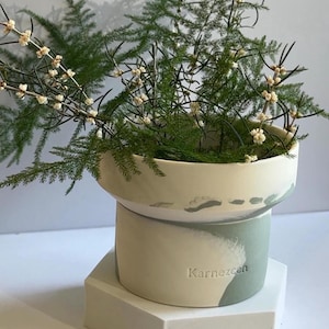 May include: A white ceramic planter with a green and white marbled design. The planter has a wide, shallow top and a narrower base. The planter is sitting on a hexagonal base. The planter has the word "Karnezcan" inscribed on the side.