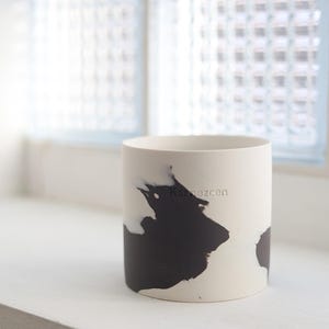 Könnte beinhalten: Ein zylindrischer Keramikbehälter mit weißem Grund und einem großen schwarzen abstrakten Design. Das Wort "Karnezcen" ist in Hellgrau auf die weiße Oberfläche gedruckt. Der Behälter steht auf einer weißen Oberfläche.