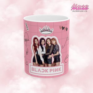 May include: A pink ceramic mug with a white interior featuring a photo of four women in black clothing. The mug has a pink grid pattern with a crown and hearts. The text "BLACKPINK" is printed in black letters on the bottom of the mug.