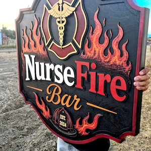 Peut inclure: Une grande enseigne en bois noir de forme personnalisée portant le texte Nurse Fire Bar et Est. 2024. Le design comprend un caducée doré dans un bouclier rouge, entouré de flammes orange et rouges en relief et d'un casque de pompier.