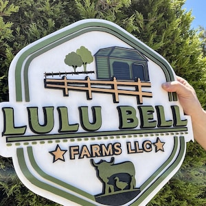 Könnte beinhalten: Ein mehrschichtiges Holzschild mit dem Text "LULU BELL FARMS LLC" in Grün und Schwarz. Das Schild zeigt eine Scheune, Bäume, einen Zaun und eine Kuh mit einer Ente. Das Schild ist weiß mit grünen und schwarzen Zierleisten.