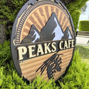 May include: A round wooden sign with the words "PEAKS CAFE" in a layered design. The sign features a mountain range graphic with a sunburst background and a black border. The sign is displayed outdoors.