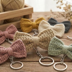 May include: A collection of handmade crochet bow tie keychains in various colors, including pink, yellow, cream, and green. Each bow tie is attached to a silver-colored keyring. The keychains are arranged on a wooden surface.
