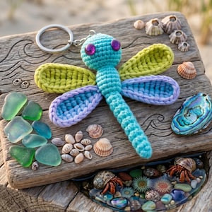 May include: A handmade crochet dragonfly keychain in shades of turquoise, green, and purple. The dragonfly is placed on a weathered wooden board with seashells, sea glass, and a decorative dish with hermit crabs.
